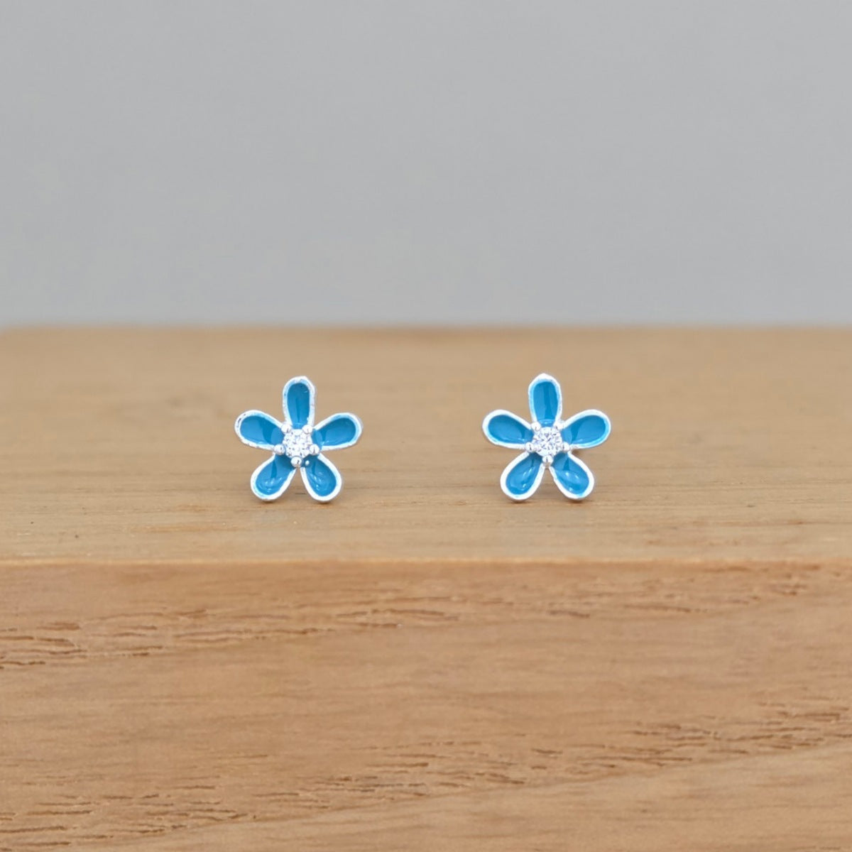 A pair of small flower-shaped stud earrings with bright blue petals and silver-toned outlines, displayed on a light wood surface against a soft gray background.