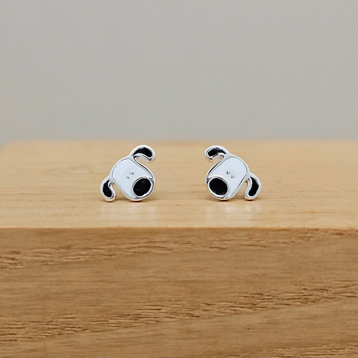 A pair of silver stud earrings shaped like cartoon dog heads with black enamel details, resting on a light wooden block against a neutral gray background.