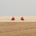 A pair of strawberry-shaped stud earrings with red and white enamel and silver-toned metal backs, displayed on a light wood block against a neutral gray background.