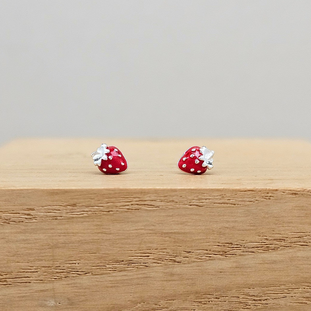 A pair of strawberry-shaped stud earrings with red and white enamel and silver-toned metal backs, displayed on a light wood block against a neutral gray background.