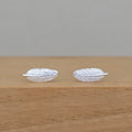 A pair of silver feather stud earrings with detailed feather textures, resting on a light brown wooden surface against a neutral gray background.