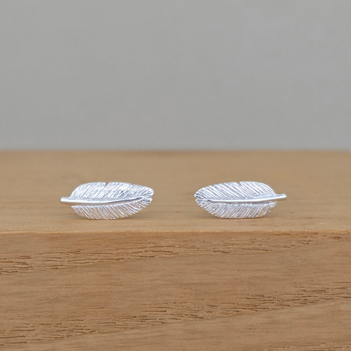 A pair of silver feather stud earrings with detailed feather textures, resting on a light brown wooden surface against a neutral gray background.