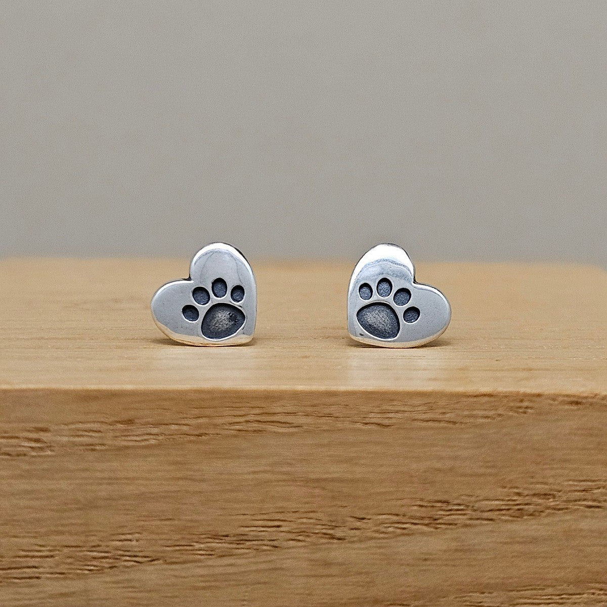 A pair of silver heart-shaped stud earrings with a dark gray paw print design in the center, displayed on a wooden block against a neutral gray background.