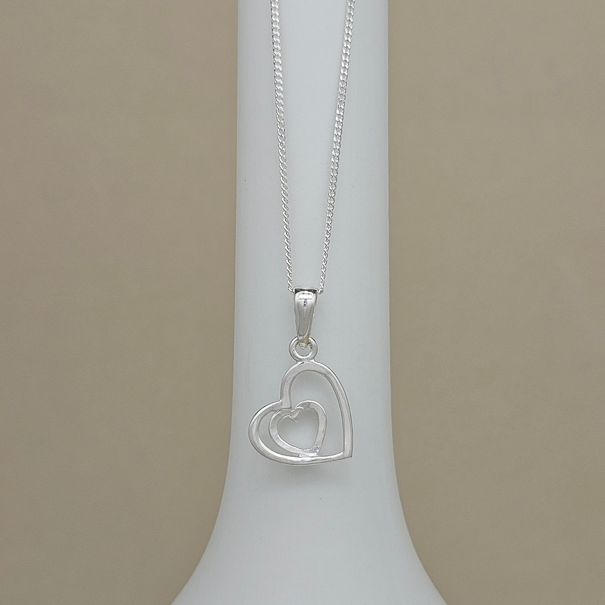 A silver necklace with a double heart pendant displayed on a white neck stand against a beige background.