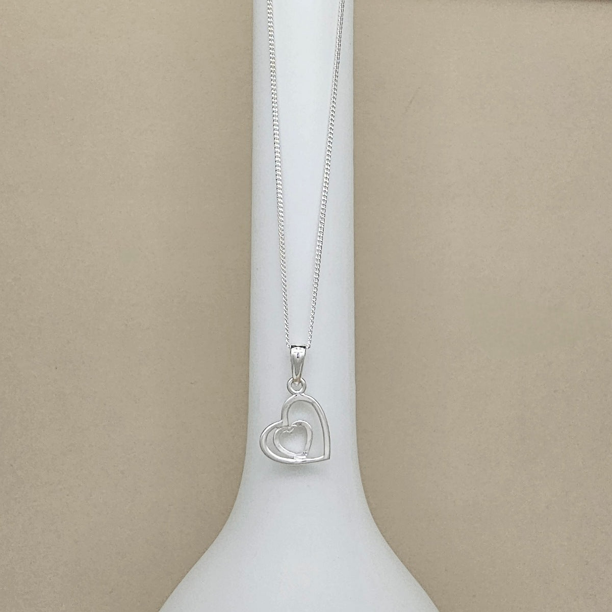 A silver necklace with a double interlocking heart pendant, displayed on a white neck mannequin against a neutral beige background.