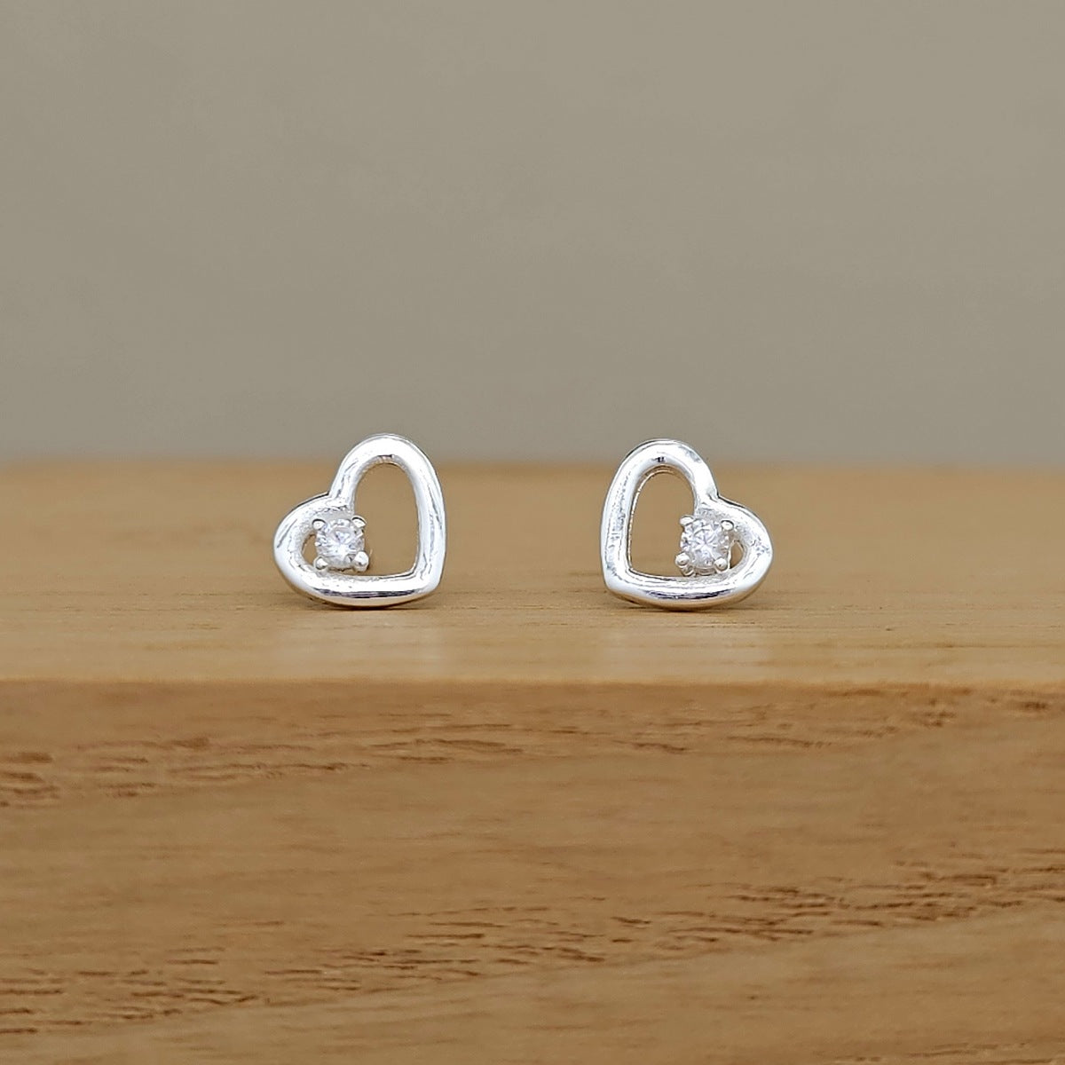 A pair of silver heart-shaped stud earrings with small clear stones, displayed on a wooden surface against a neutral beige background.