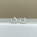 A pair of silver horseshoe-shaped stud earrings with small clear stones set along the outer curve, displayed on a white surface against a beige background.
