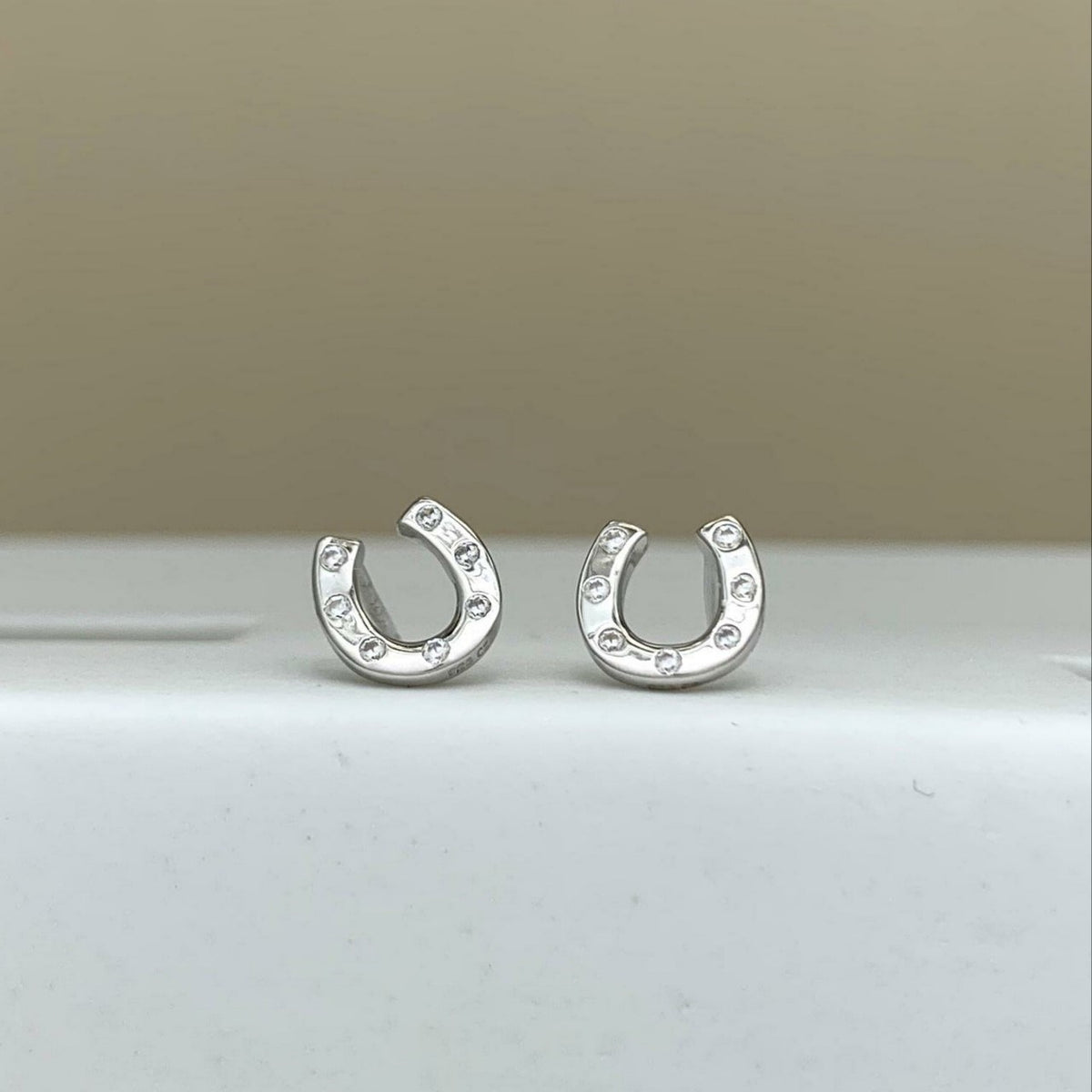 A pair of silver horseshoe-shaped stud earrings with small clear stones set along the outer curve, displayed on a white surface against a beige background.