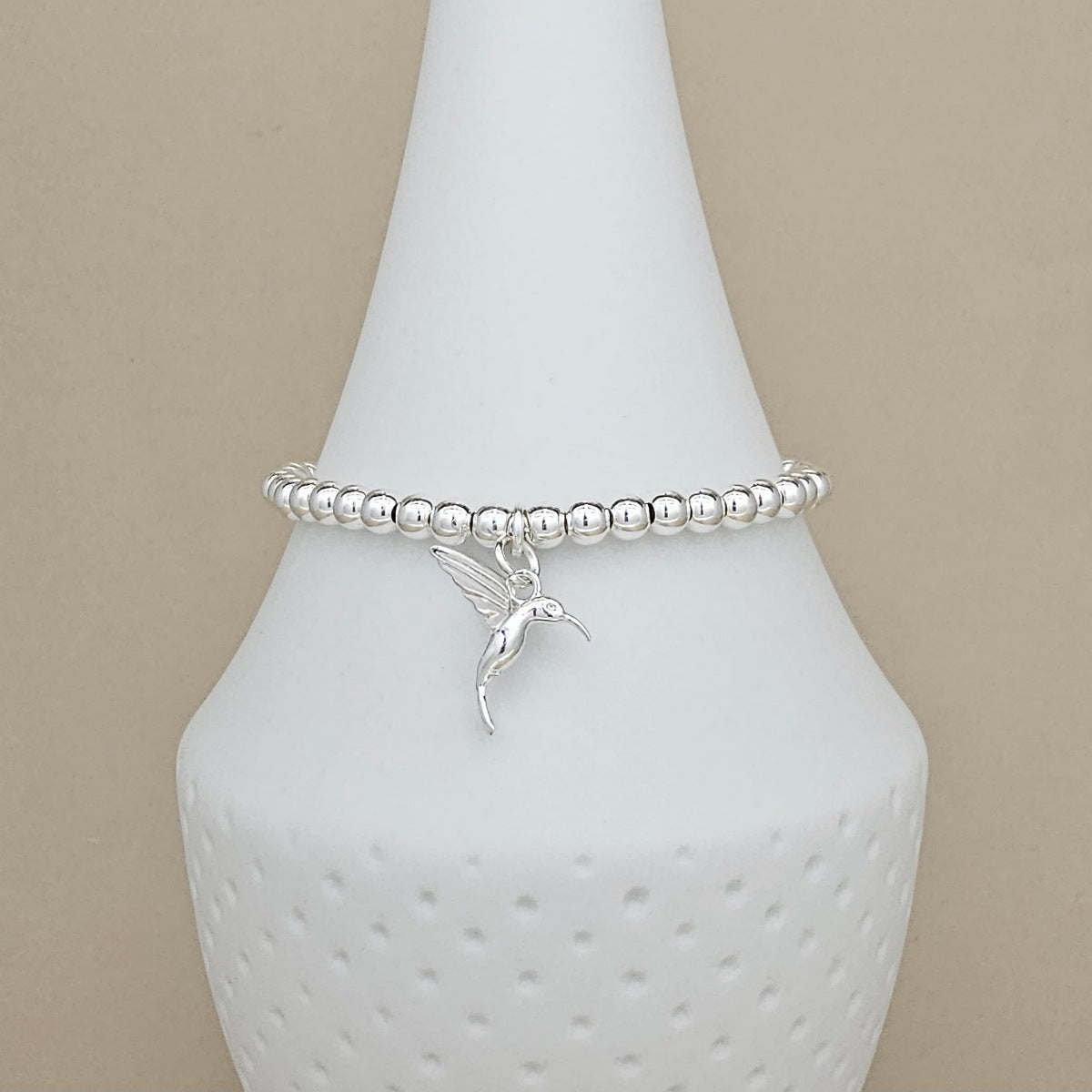 A silver beaded bracelet with a small hummingbird charm, displayed on a white ceramic jewelry bust against a neutral beige background.