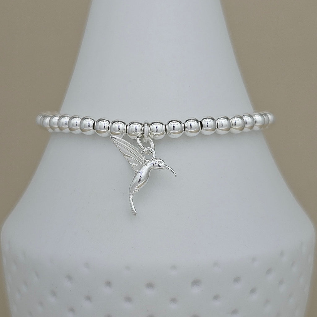 A silver beaded bracelet with a small hummingbird charm, displayed on a white cone jewelry stand against a neutral beige background.