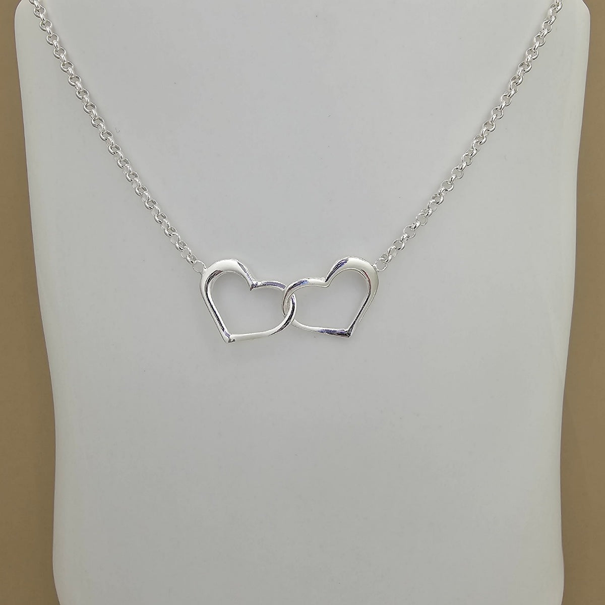 A silver chain necklace with two interlocking open heart pendants, displayed on a white mannequin bust against a beige background.