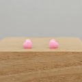 A pair of small, glossy pink heart-shaped stud earrings resting on a light brown wooden block against a neutral gray background.