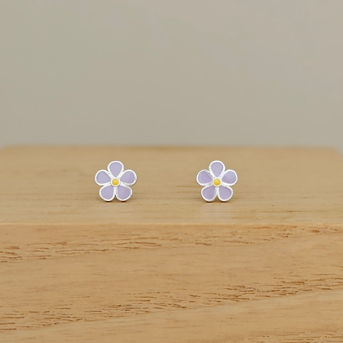 A pair of small flower-shaped stud earrings with lavender-purple petals, white outlines, and yellow centers, displayed on a light wood surface against a neutral beige background.