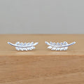 A pair of silver leaf-shaped stud earrings resting on a light brown wooden surface against a neutral gray background.