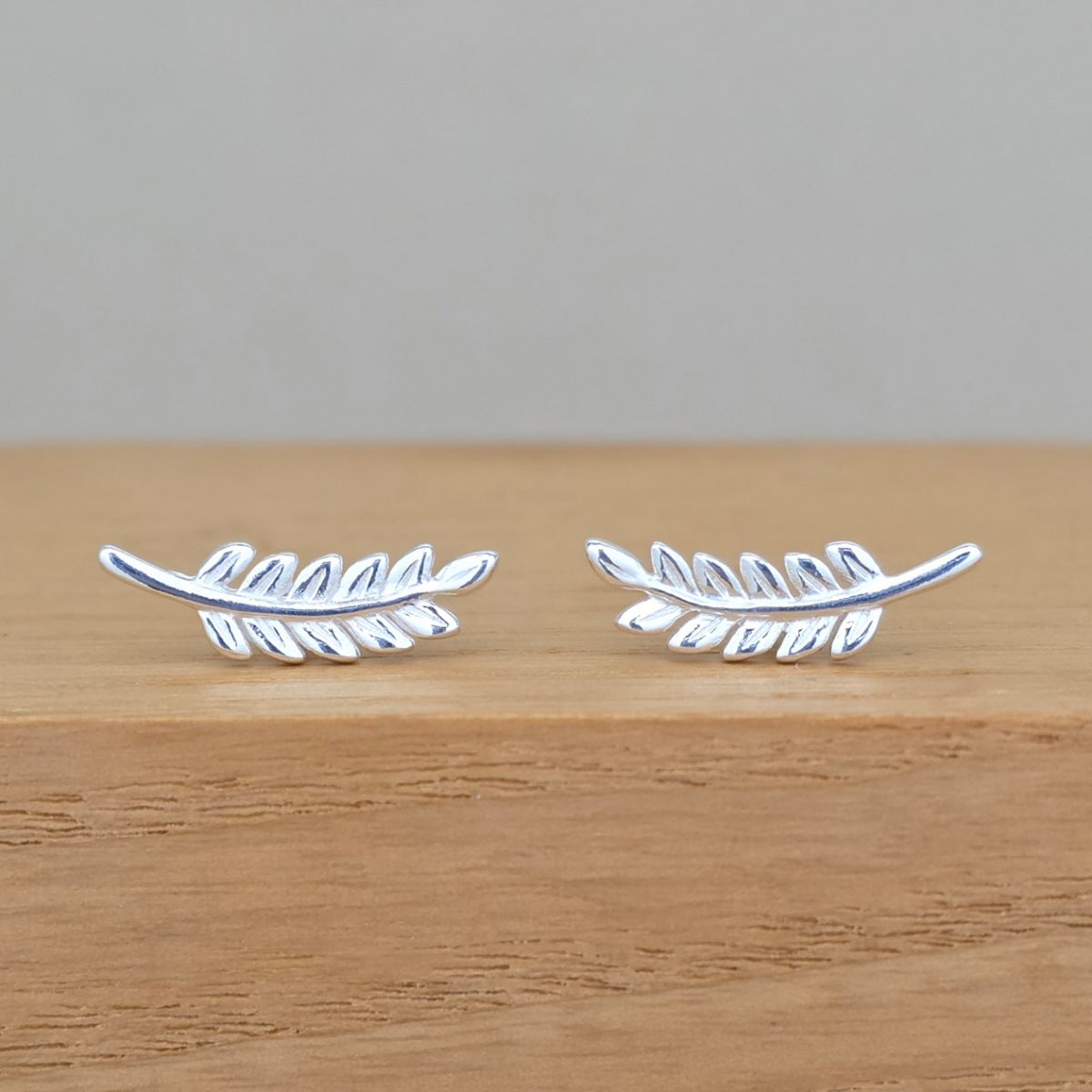 A pair of silver leaf-shaped stud earrings resting on a light brown wooden surface against a neutral gray background.