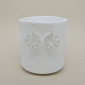 A pair of silver circular dangle earrings with delicate leaf cutout details, displayed on a matte white ceramic cup against a warm beige background.