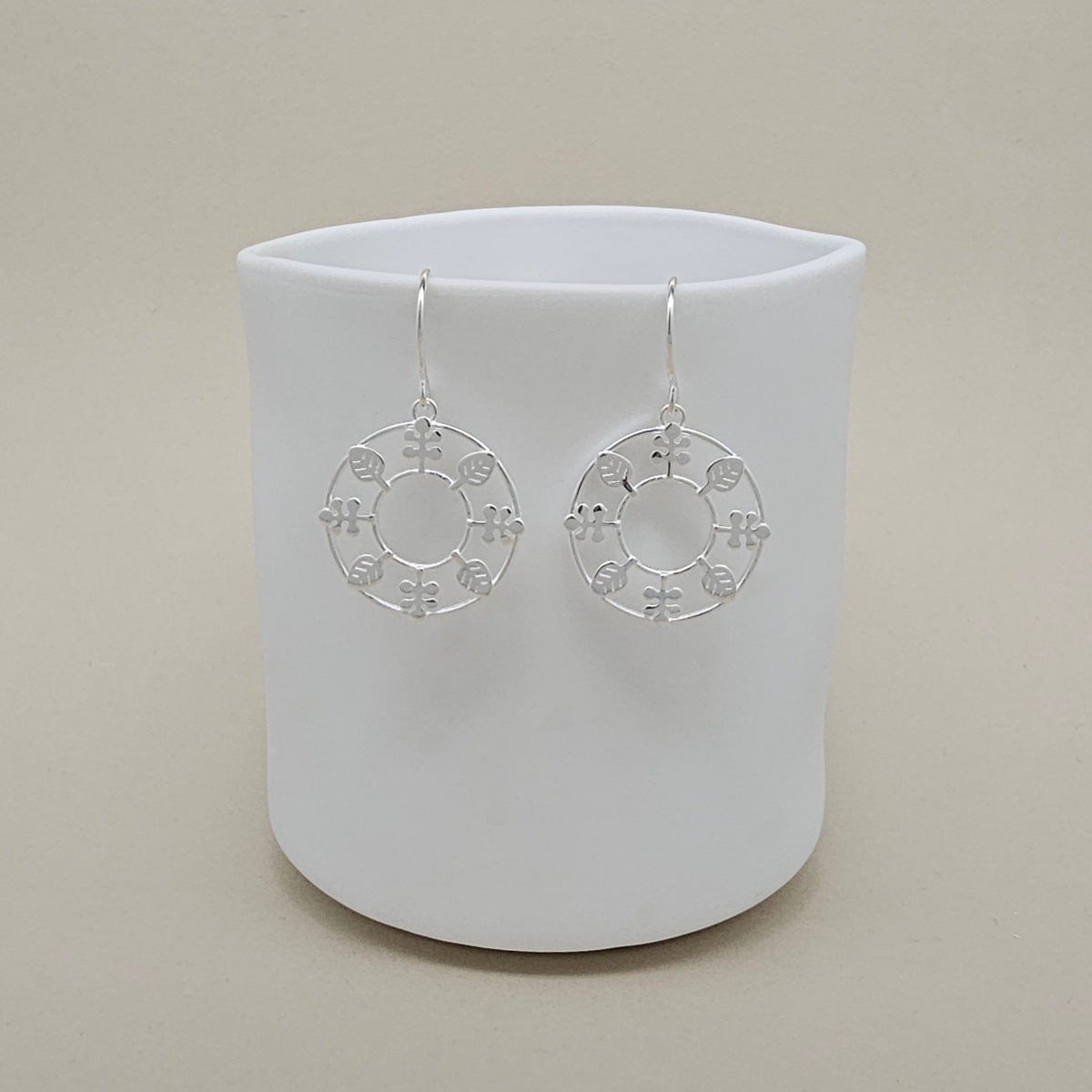 A pair of silver circular dangle earrings with delicate leaf cutout details, displayed on a matte white ceramic cup against a warm beige background.