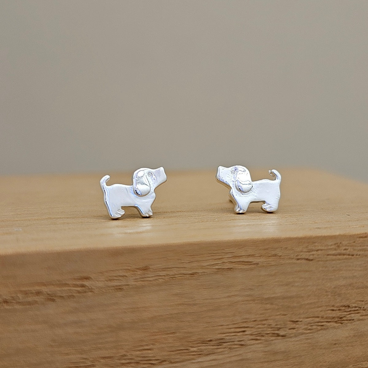 A pair of small silver stud earrings shaped like dogs, displayed on a light wooden block against a neutral beige background.