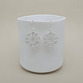 A pair of silver circular dangle earrings with cutout leaves and heart patterns, hanging from a white ceramic cup against a warm beige background.