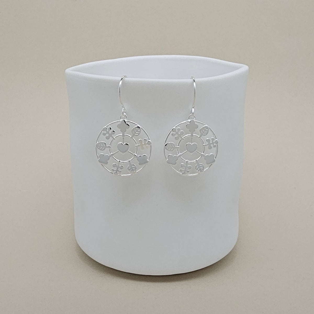 A pair of silver circular dangle earrings with cutout leaves and heart patterns, hanging from a white ceramic cup against a warm beige background.