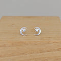 A pair of silver crescent moon and star stud earrings displayed on a light wood surface against a neutral gray background.
