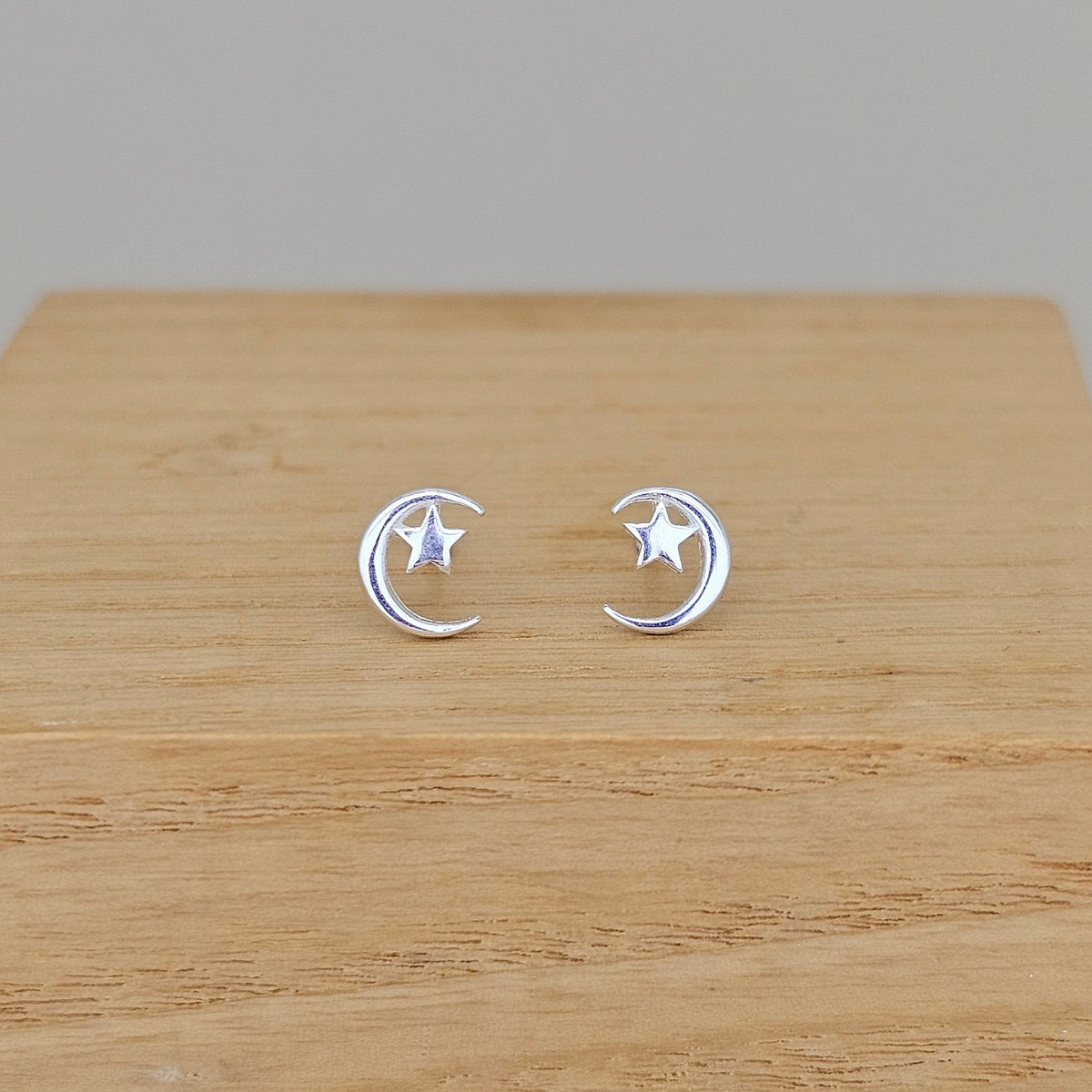 A pair of silver crescent moon and star stud earrings displayed on a light wood surface against a neutral gray background.