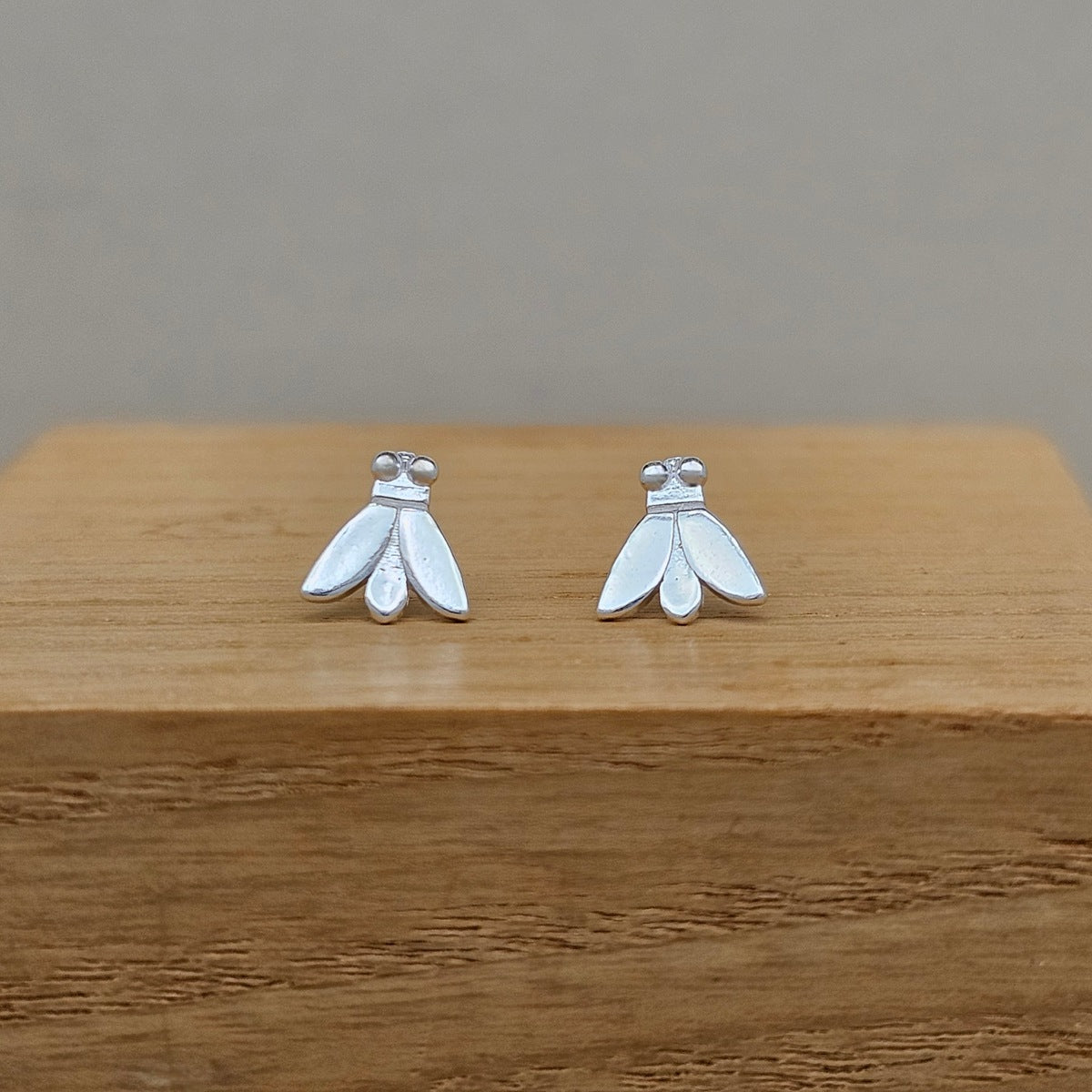 A pair of small silver moth stud earrings displayed on a wooden block against a neutral gray background.