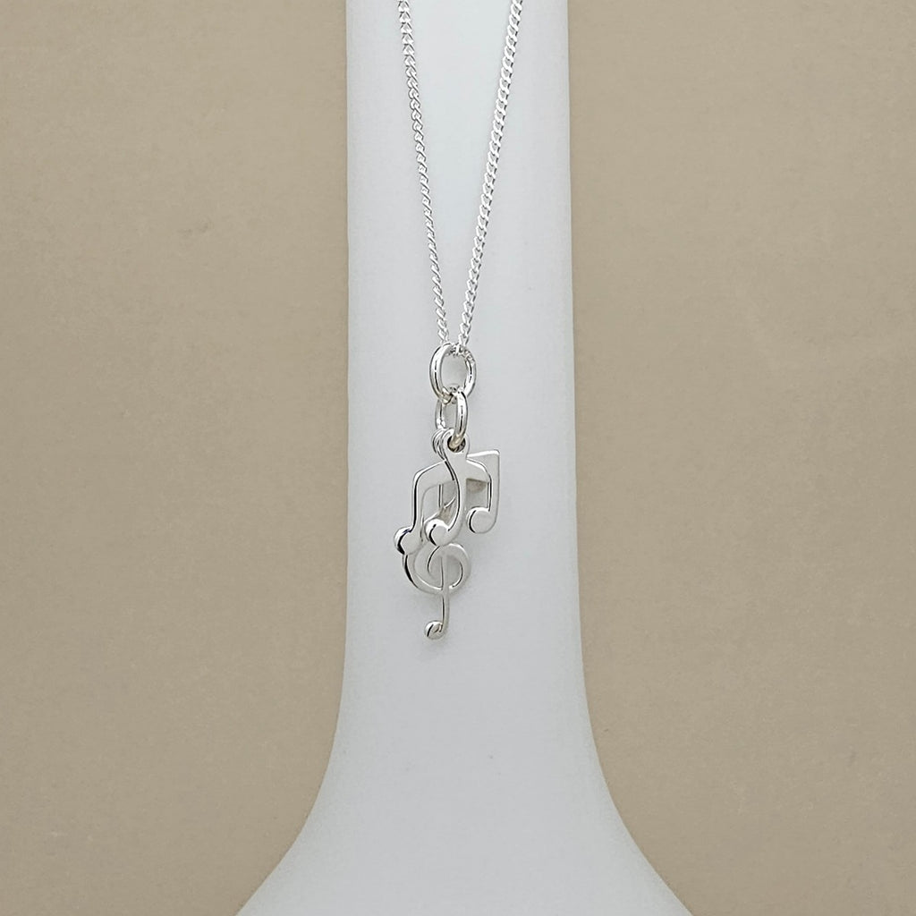 A silver necklace with a musical note and treble clef pendant, displayed on a white neck stand against a beige background.