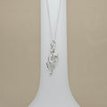 A silver necklace with a musical note and treble clef pendant, displayed on a white neck stand against a beige background.