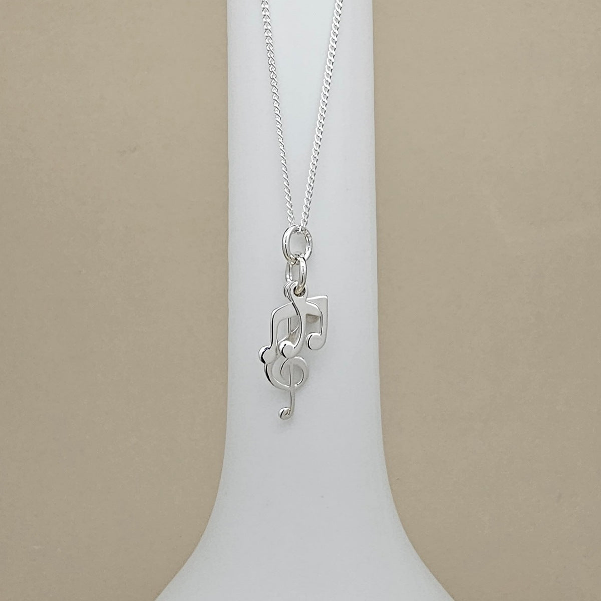 A silver necklace with a musical note and treble clef pendant, displayed on a white neck stand against a beige background.