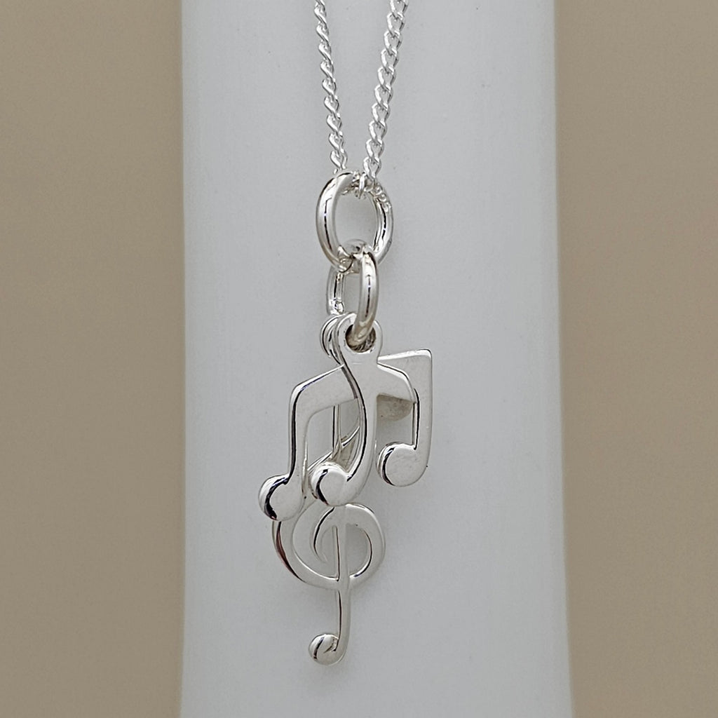 A silver necklace with a pendant shaped like a treble clef intertwined with musical notes, displayed on a white cylindrical stand against a beige background.