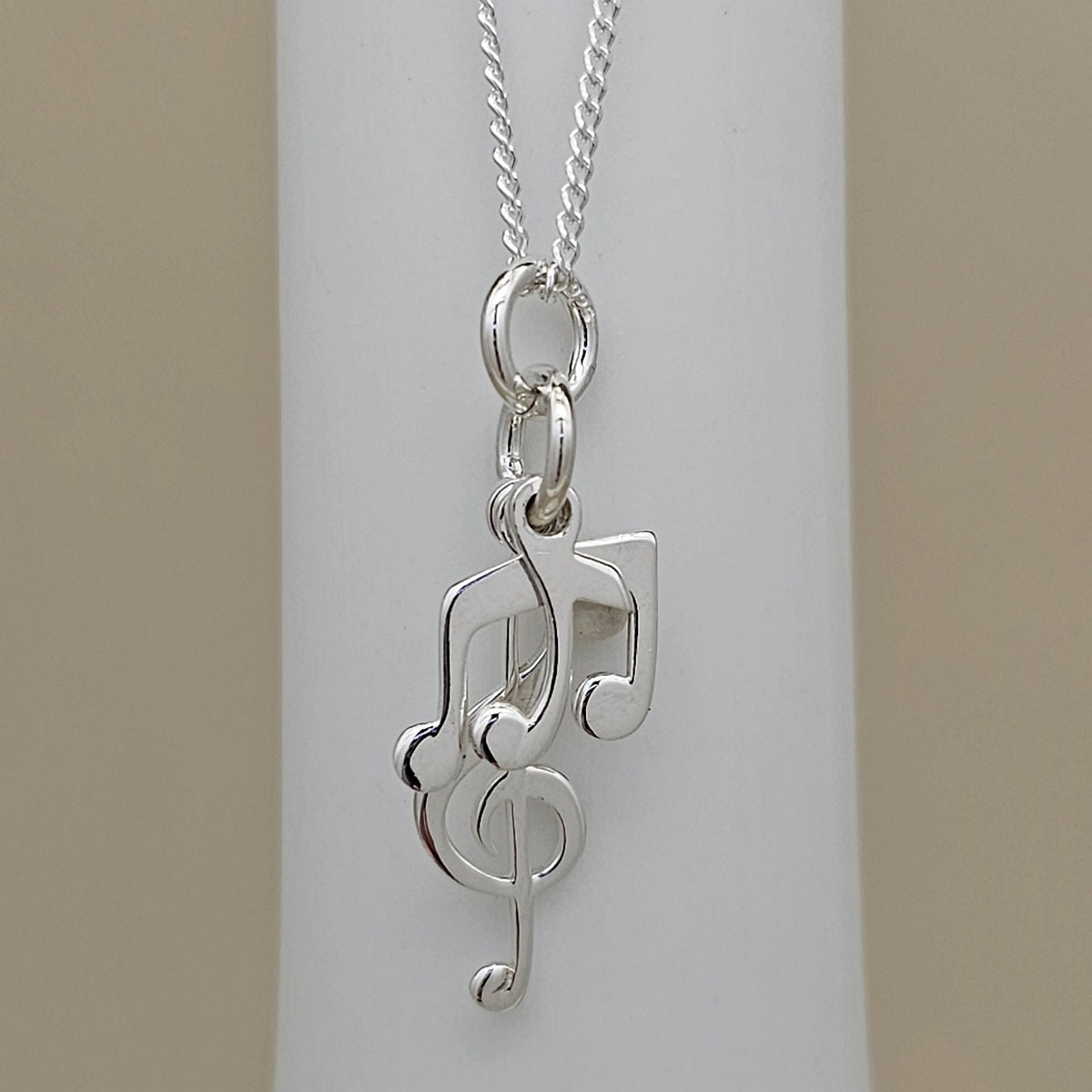 A silver necklace with a pendant shaped like a treble clef intertwined with musical notes, displayed on a white cylindrical stand against a beige background.