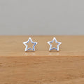 A pair of silver star-shaped stud earrings displayed on a wooden surface against a neutral gray background.