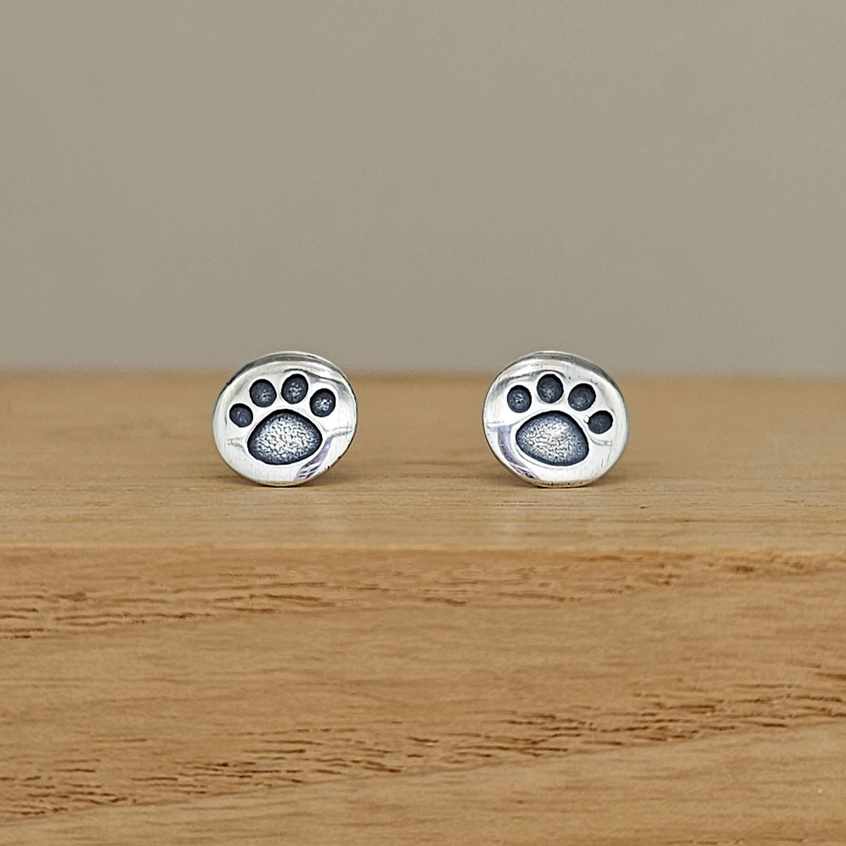 A pair of silver stud earrings shaped like paw prints, displayed on a light wooden surface against a neutral beige background.