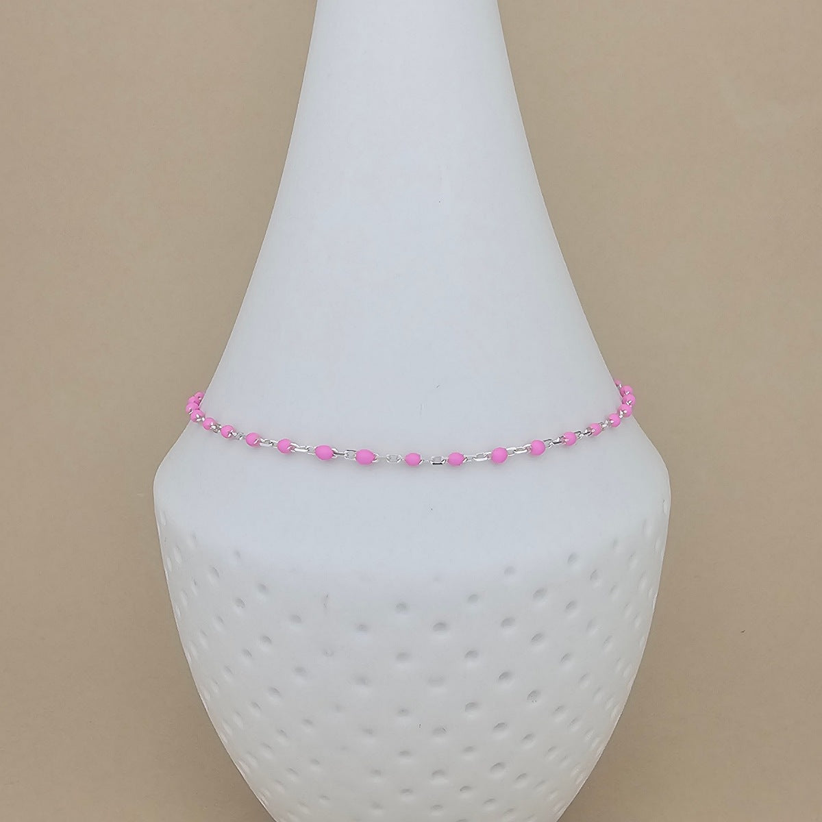 A delicate beaded anklet with pink and silver beads displayed on a white cone-shaped jewelry stand against a solid beige background.