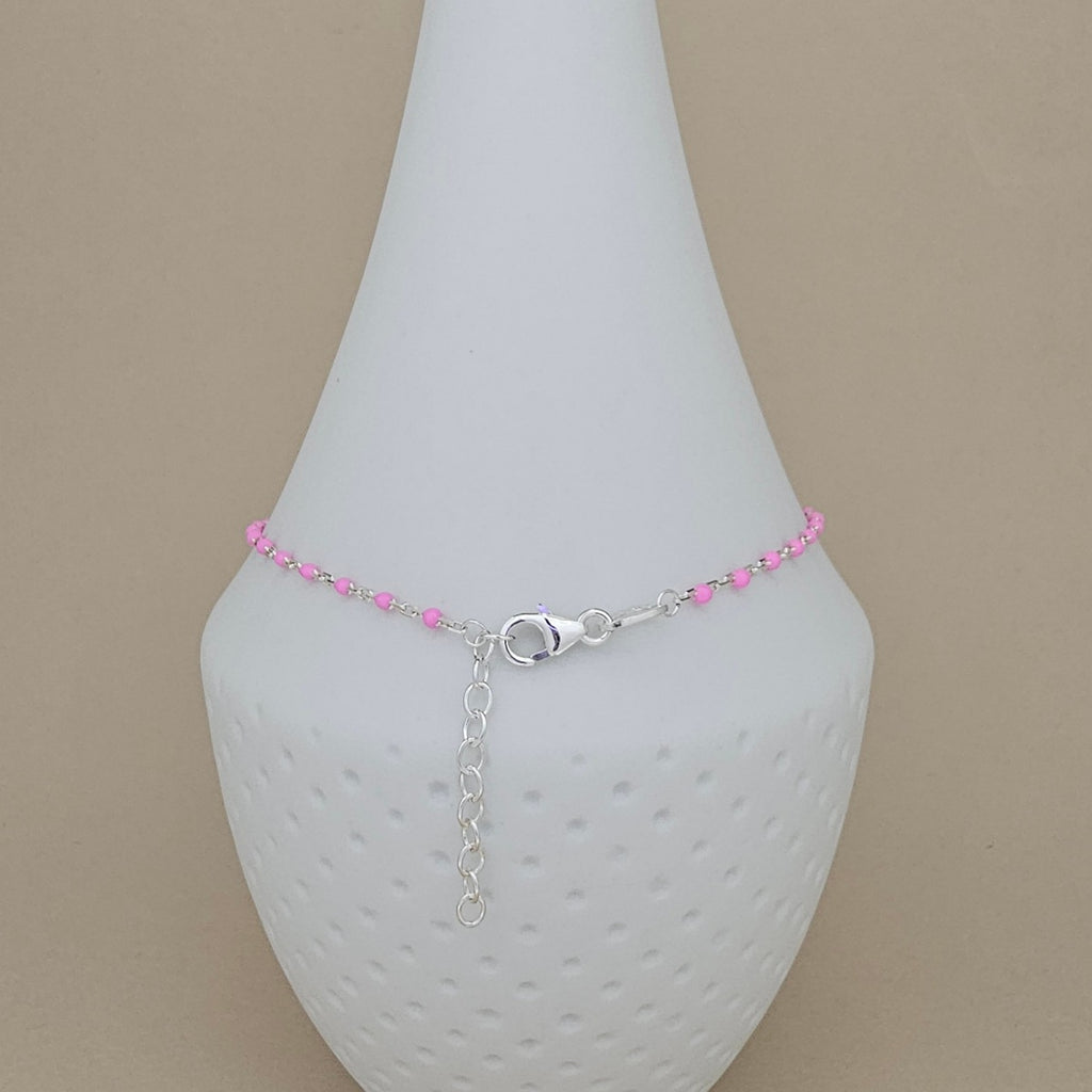 A delicate silver chain anklet with small pink beads and a silver lobster clasp, displayed on a white textured cone stand against a beige background.