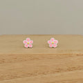 A pair of small flower-shaped stud earrings with pink petals, silver outlines, and yellow centers, displayed on a light wood surface against a neutral beige background.