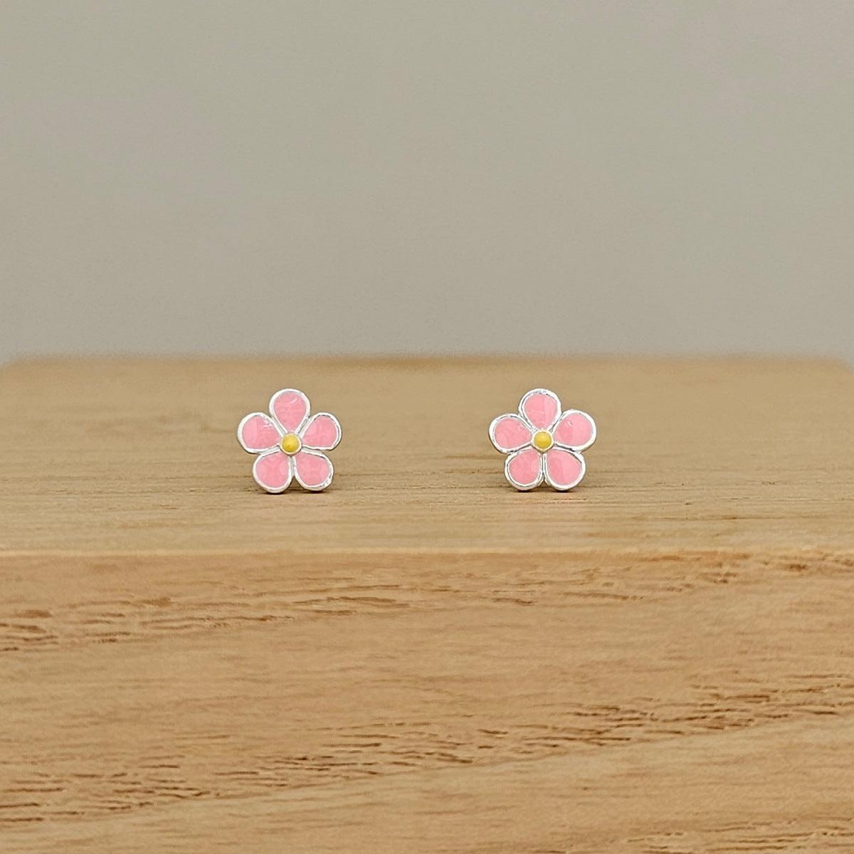 A pair of small flower-shaped stud earrings with pink petals, silver outlines, and yellow centers, displayed on a light wood surface against a neutral beige background.