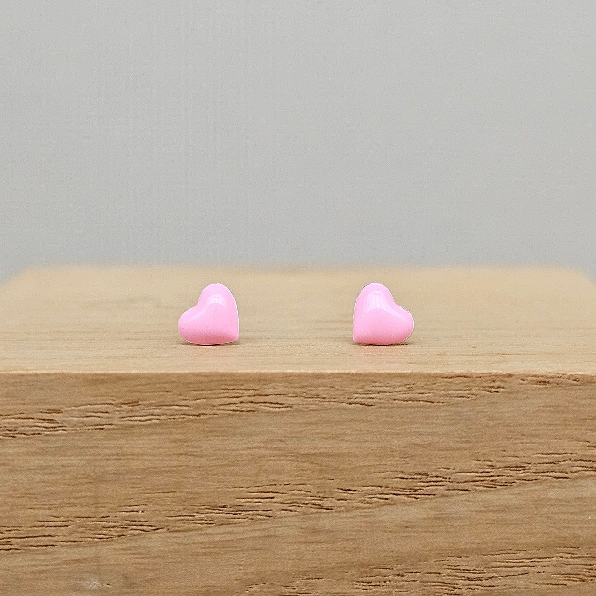 A pair of small pink heart-shaped stud earrings resting on a light wood block against a plain light gray background.