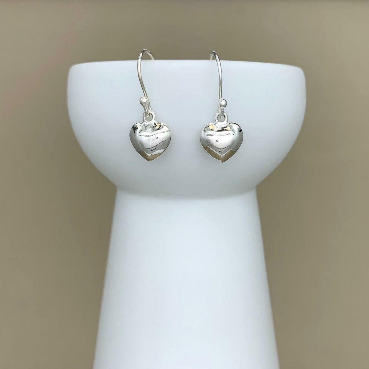 A pair of polished silver heart-shaped drop earrings hanging from silver fishhook wires, displayed on a white ceramic jewelry stand against a warm beige background.