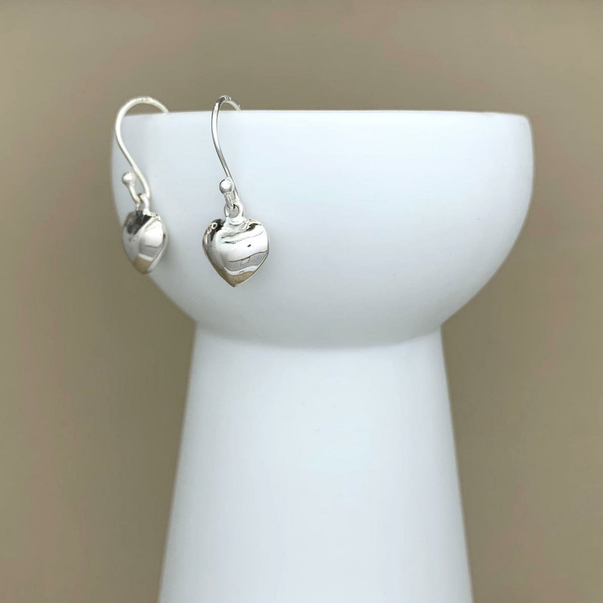 A pair of polished silver heart-shaped dangle earrings displayed on a white ceramic jewelry stand against a soft beige background.