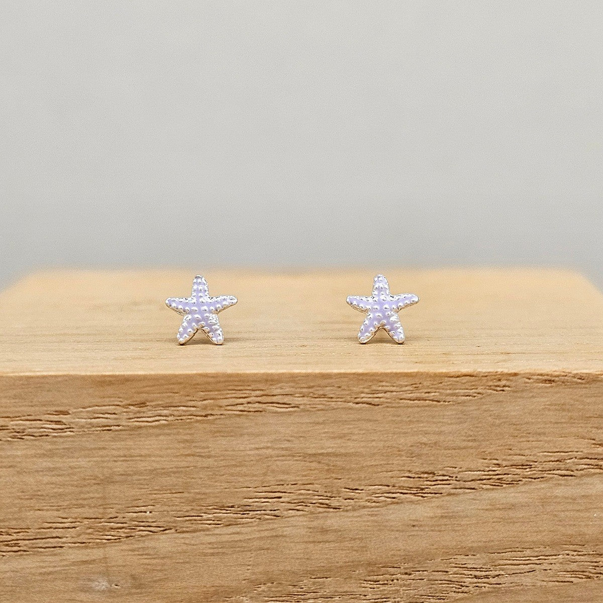 A pair of small starfish-shaped stud earrings with purple enamel, displayed on a light wood block against a soft gray background.