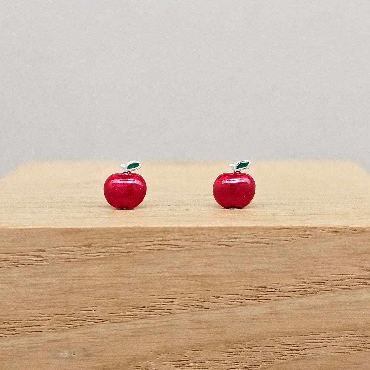 Two red apple-shaped earrings on a wooden surface with a gray background