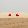 A pair of small, glossy red heart-shaped stud earrings resting on a light-colored wooden block against a neutral gray background.