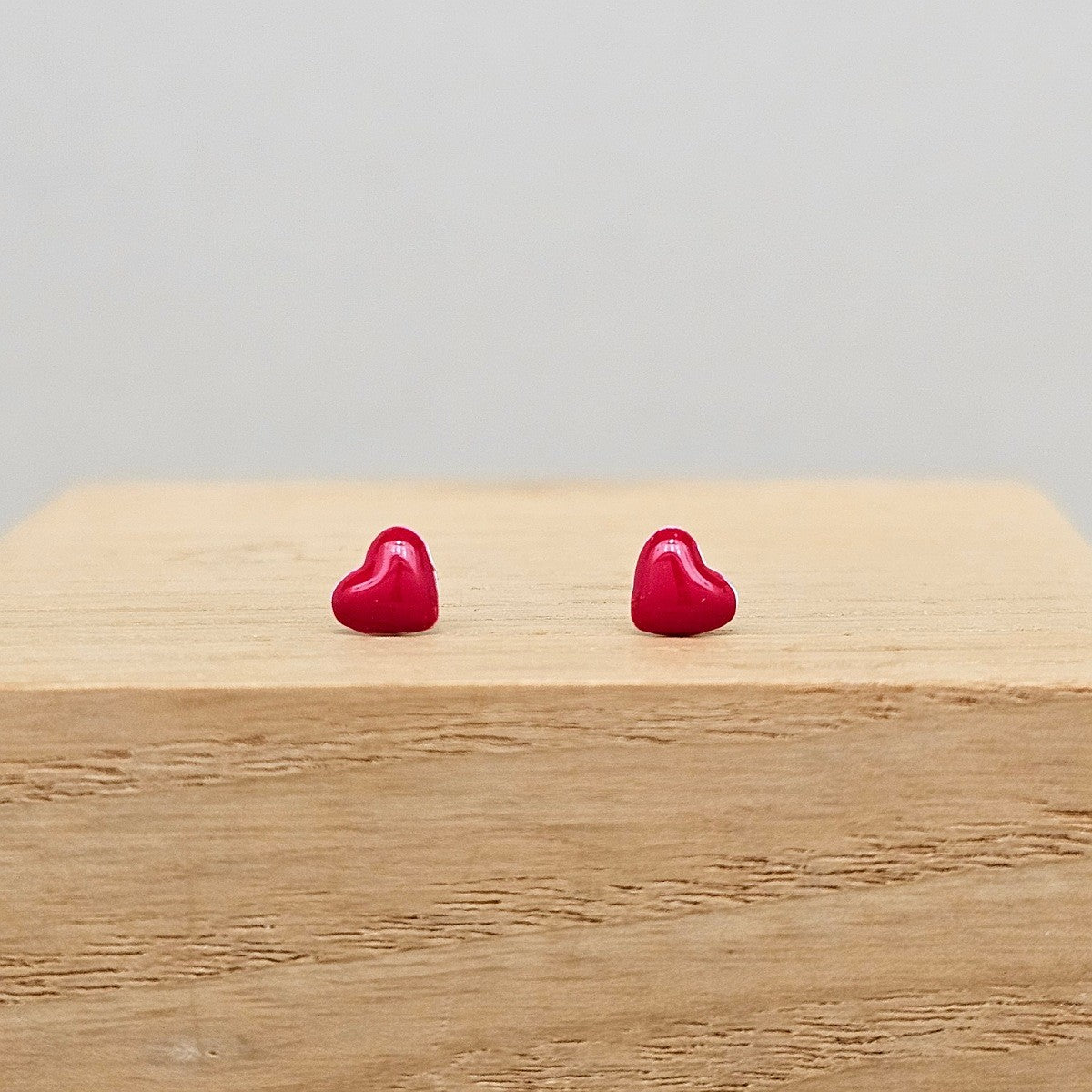 A pair of small, glossy red heart-shaped stud earrings resting on a light-colored wooden block against a neutral gray background.