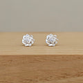 A pair of silver rose stud earrings with clear central gemstones, resting on a light wood surface against a neutral beige background.