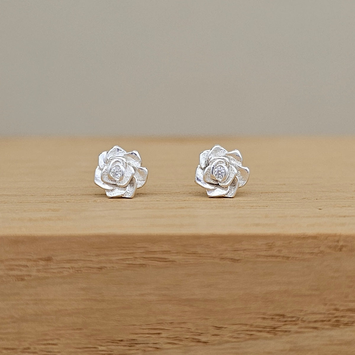 A pair of silver rose stud earrings with clear central gemstones, resting on a light wood surface against a neutral beige background.
