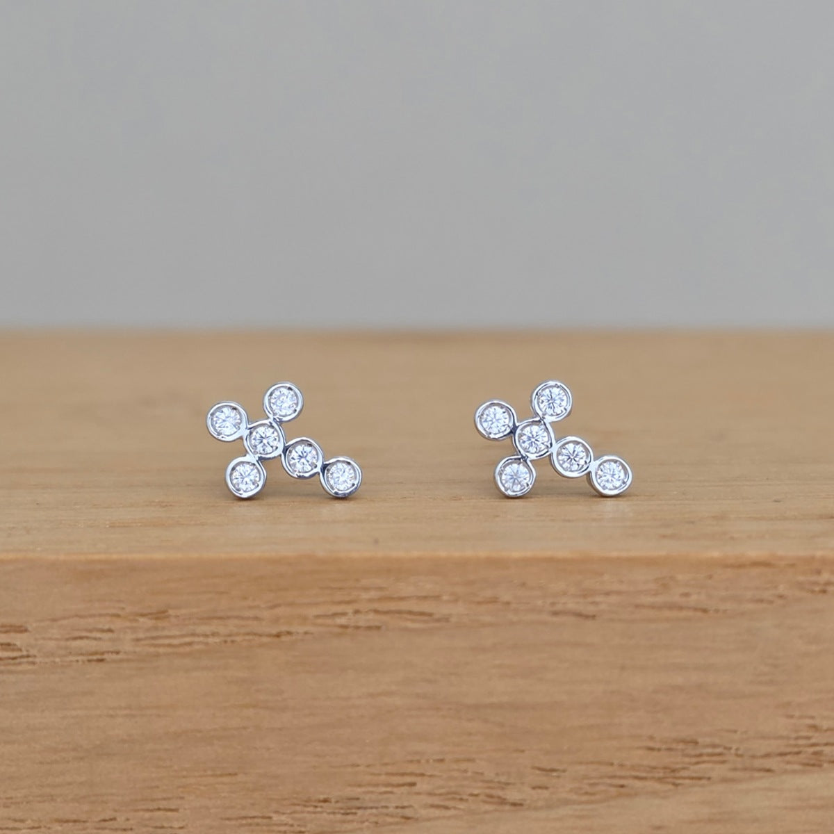 A pair of silver stud earrings with five clear round-cut stones arranged in a small cluster, resting on a light brown wooden surface against a neutral gray background.
