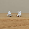 A pair of silver squirrel stud earrings resting on a light brown wooden surface against a neutral beige background.