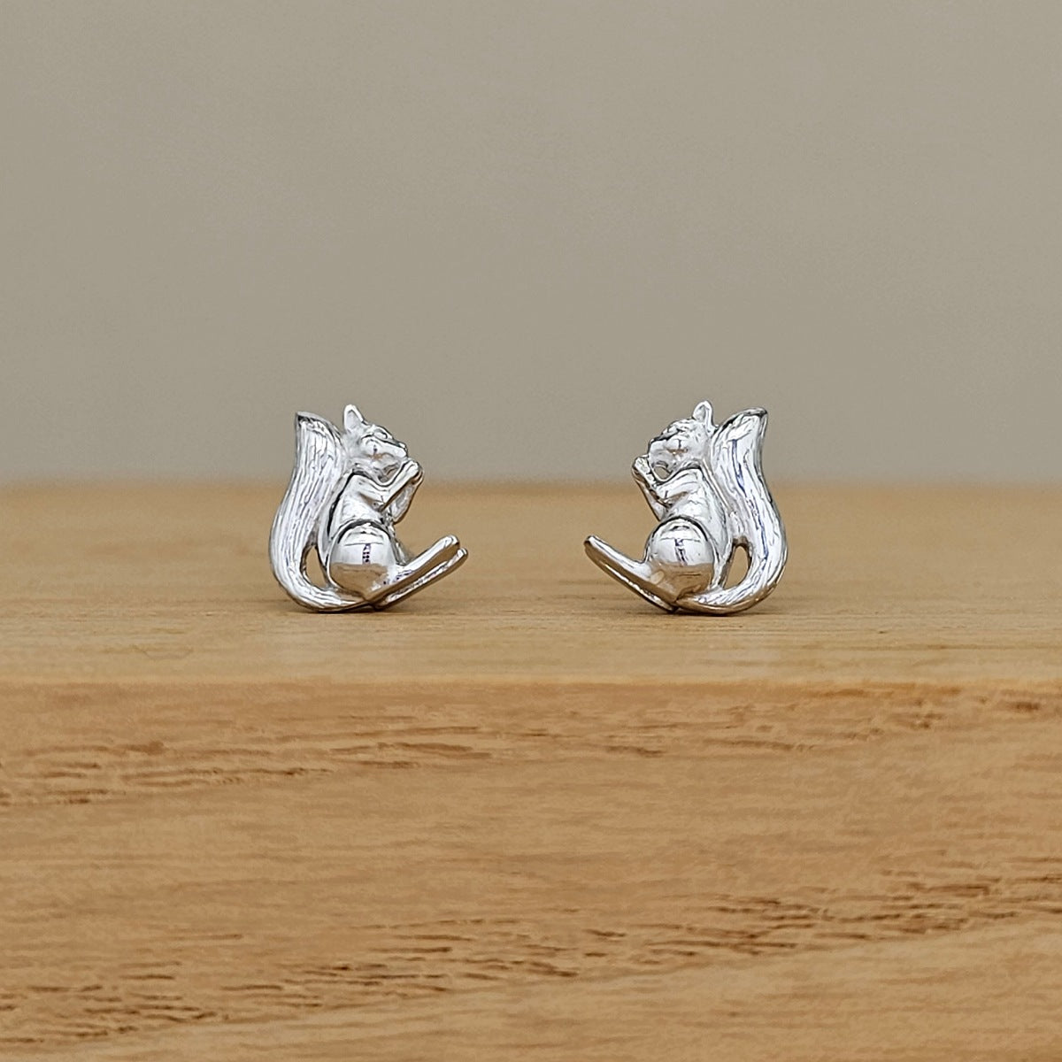 A pair of silver squirrel stud earrings resting on a light brown wooden surface against a neutral beige background.